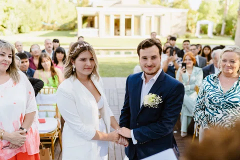 Mujer vestida de blanco y hombre con traje azul están de pie tomados de la mano, con personas sentadas detrás.