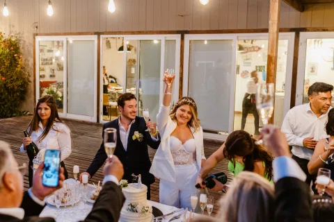 Mujer vestida de blanco levanta una mano junto a una mesa rodeada por varias personas.