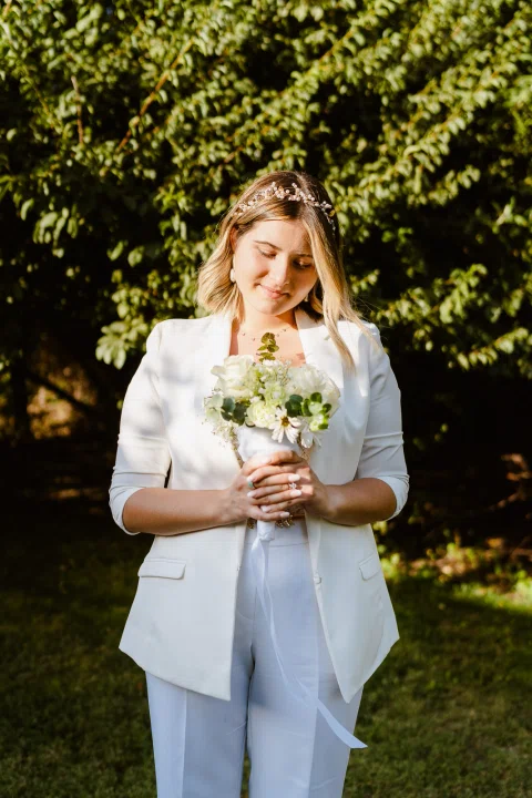 Mujer vestida de blanco posa de pie con un ramo de flores blancas en un jardín.