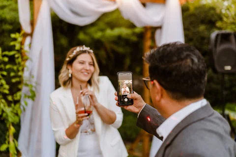 Mujer vestida de blanco sostiene una copa mientras un hombre en primer plano la fotografía con un teléfono celular.