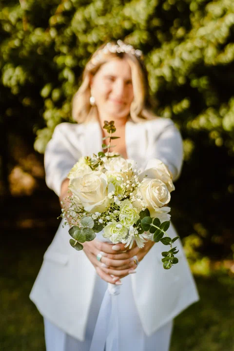 Mujer vestida de blanco sostiene un ramo de flores blancas hacia la cámara en un jardín.