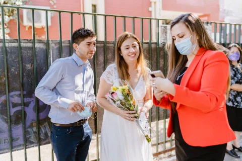 Mujer con blazer rojo mira la pantalla de un teléfono junto a un hombre con camisa celeste y una mujer con vestido blanco y ramo.