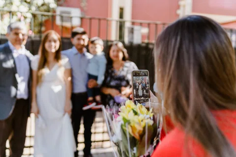 Mujer con blazer rojo sostiene un teléfono en primer plano frente a un grupo de personas en el exterior.