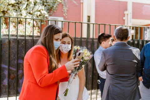 Mujer con blazer rojo y mujer con vestido blanco y ramo miran juntas la pantalla de un teléfono en el exterior.