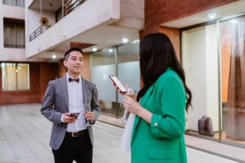 Mujer con blazer verde consulta un celular mientras conversa con un hombre con traje gris que sostiene una copa.