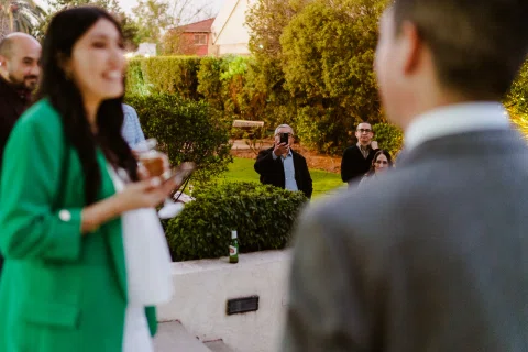 Mujer con blazer verde conversa frente a un hombre con traje gris mientras varias personas permanecen al fondo.