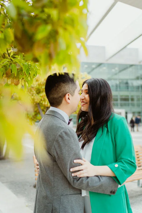 Mujer con blazer verde y hombre con traje gris se besan junto a un árbol.