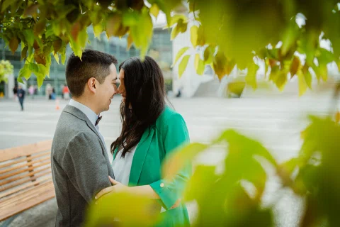 Mujer con blazer verde y hombre con traje gris se besan bajo hojas verdes desenfocadas.