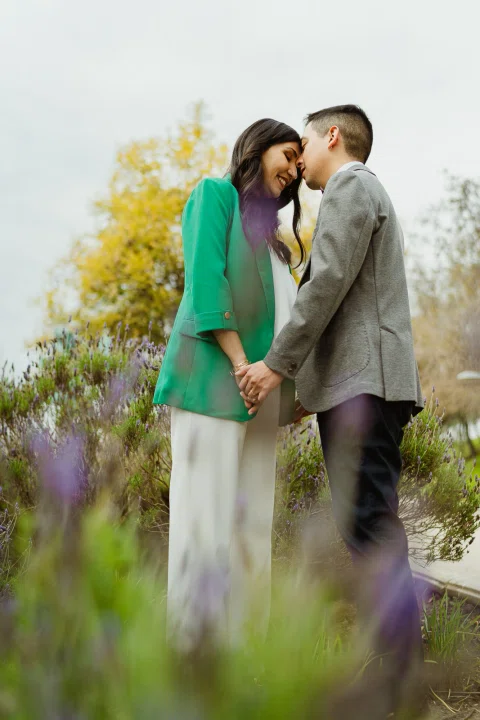 Mujer con blazer verde y hombre con traje gris se besan detrás de flores lilas desenfocadas.