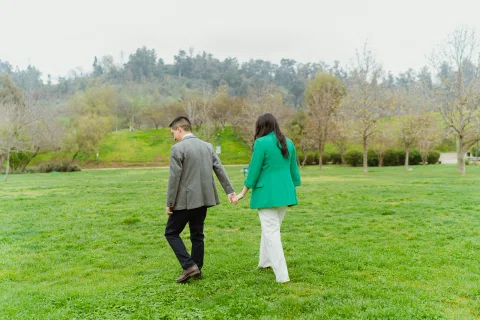 Mujer con blazer verde y hombre con traje gris caminan de espaldas tomados de la mano sobre el césped.