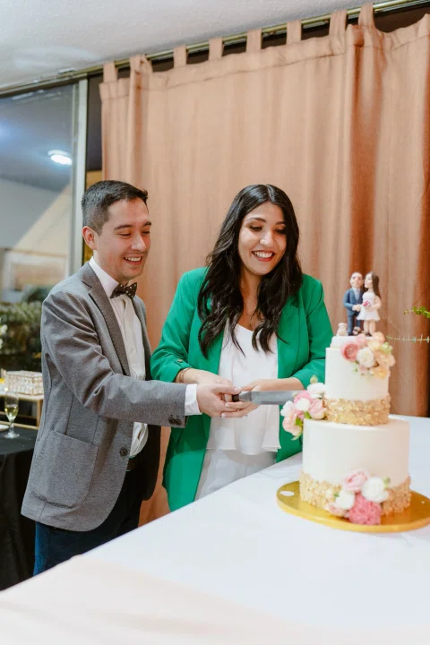 Mujer con blazer verde y hombre con traje gris cortan juntos una torta decorada.