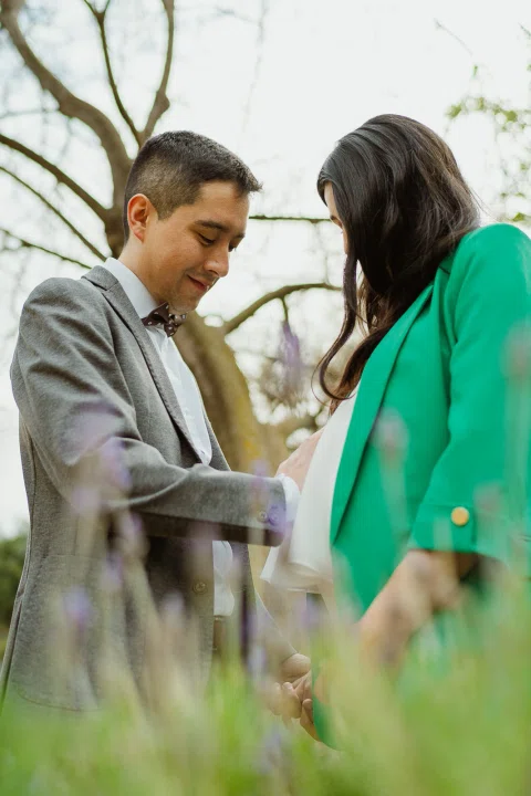 Mujer con blazer verde y hombre con traje gris se miran detrás de vegetación desenfocada.