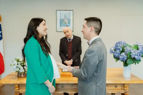 Mujer con blazer verde y hombre con traje gris permanecen frente a frente tomados de las manos, con un hombre mayor detrás.