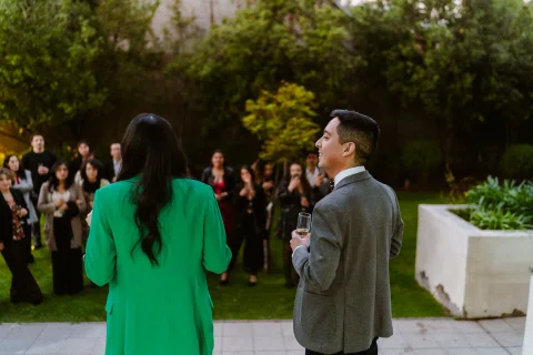 Mujer con blazer verde y hombre con traje gris permanecen frente a un grupo reunido en un patio exterior.