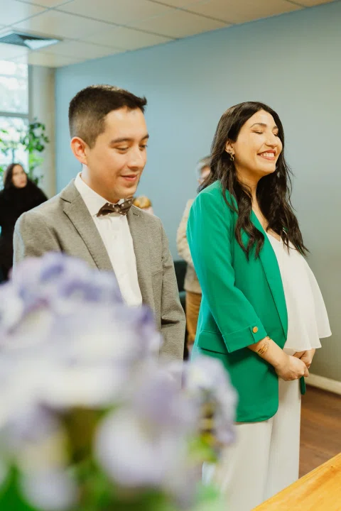 Mujer con blazer verde y hombre con traje gris permanecen frente a un hombre mayor detrás de una mesa, con flores desenfocadas en primer plano.