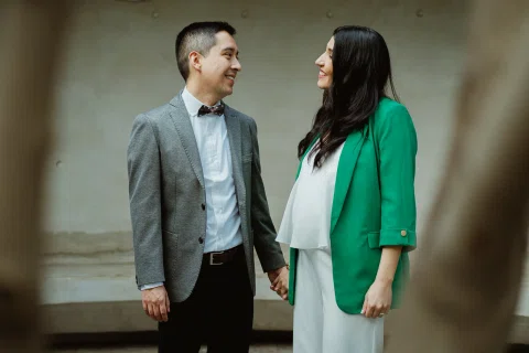 Mujer con blazer verde y hombre con traje gris permanecen tomados de la mano frente a un muro liso.