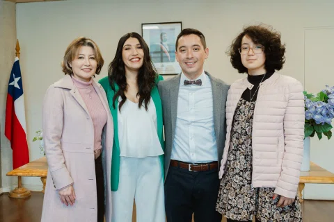 Mujer con blazer verde y hombre con traje gris posan con dos mujeres frente a una bandera chilena.