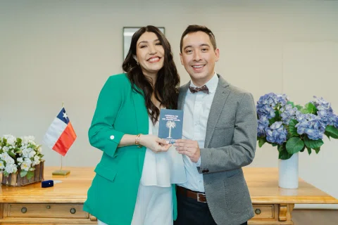 Mujer con blazer verde y hombre con traje gris sonríen mientras sostienen juntos un pequeño cuaderno morado.