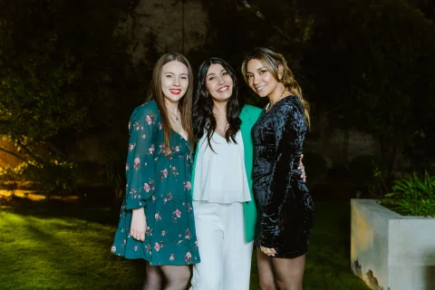 Mujer con blazer verde posa junto a dos mujeres en un jardín de noche.