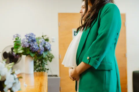 Mujer con blazer verde posa de perfil con las manos sobre el abdomen junto a un arreglo de flores lilas.