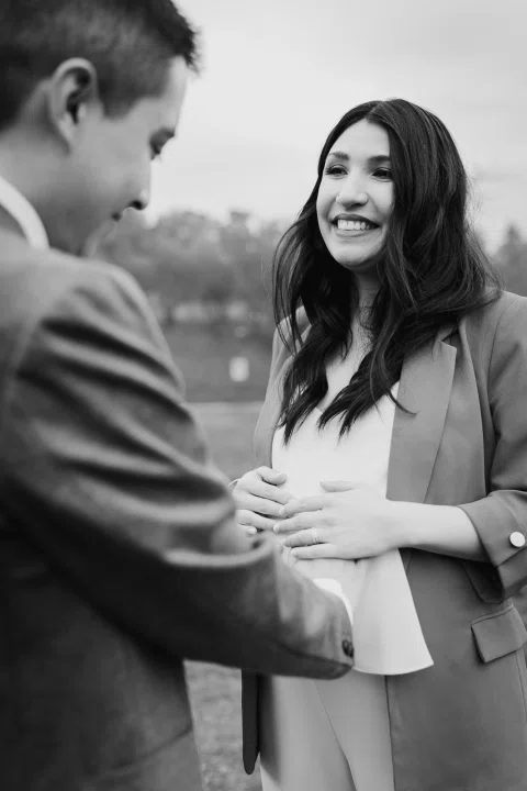 Mujer con blazer verde sonríe hacia un hombre con traje gris, en blanco y negro.