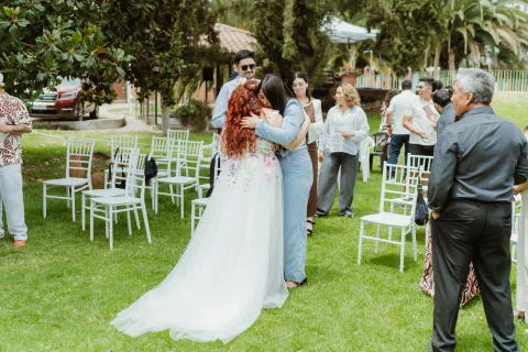 Mujer de cabello rojo abraza a una mujer con vestido celeste entre sillas blancas.