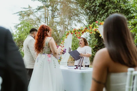 Mujer de cabello rojo con vestido floral permanece frente a una mujer con vestido blanco junto a una mesa.
