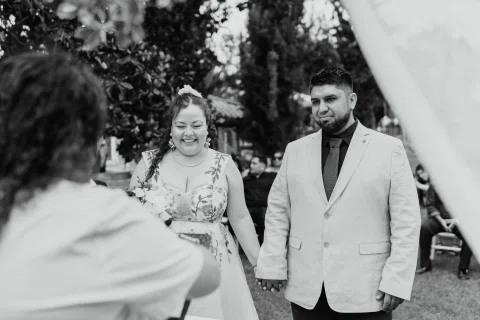 Mujer de cabello rojo con ramo y hombre con chaqueta clara permanecen juntos frente a una mujer de espaldas, en una fotografía en blanco y negro.