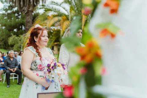 Mujer de cabello rojo sostiene un ramo y sonríe junto a un arco floral.