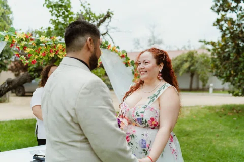 Mujer de cabello rojo sonríe frente a un hombre con chaqueta clara junto a un arco floral.