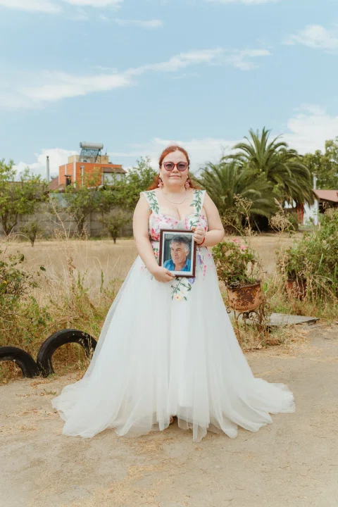 Mujer de cabello rojo con vestido floral y falda blanca posa al aire libre sosteniendo una fotografía enmarcada.