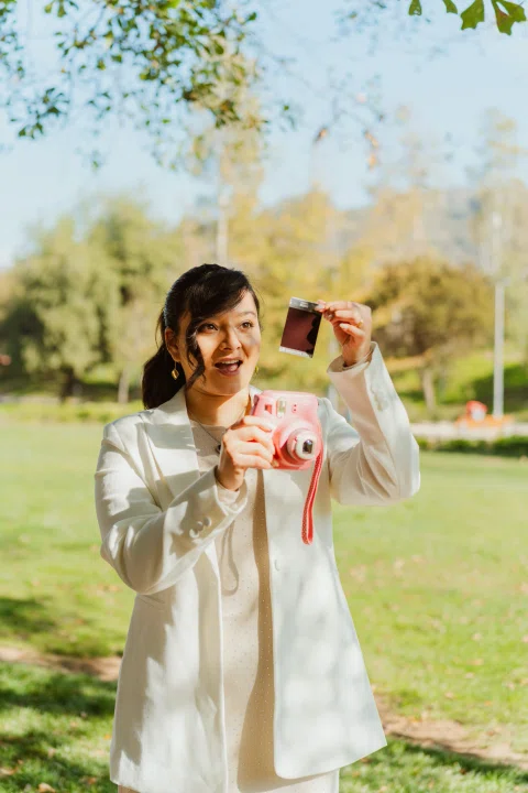 Mujer con traje claro sostiene una cámara instantánea rosada y muestra una fotografía.