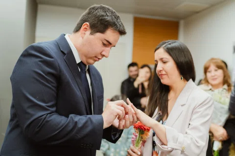 Mujer de chaqueta clara coloca un anillo en el dedo de un hombre con traje oscuro.