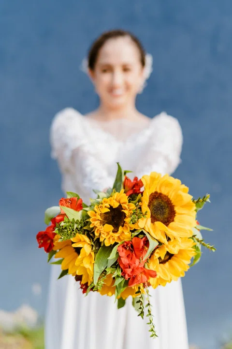 Mujer posando de frente con un ramo de girasoles y flores naranjas.
