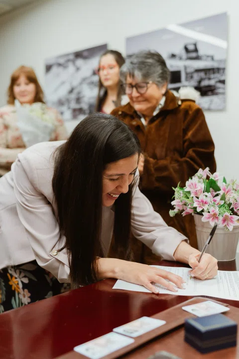 Mujer de chaqueta clara firma un documento sobre una mesa.