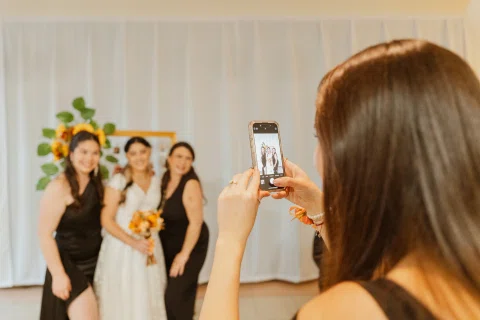 Mujer de espaldas fotografía con un teléfono a tres mujeres, una de ellas con vestido blanco y ramo.