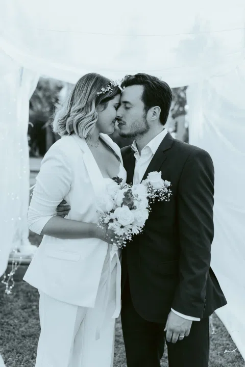 Mujer vestida de blanco y hombre con traje oscuro se besan con un ramo en primer plano en una fotografía en blanco y negro.