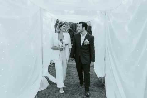Mujer vestida de blanco y hombre con traje oscuro caminan tomados de la mano entre telas blancas en una fotografía en blanco y negro.