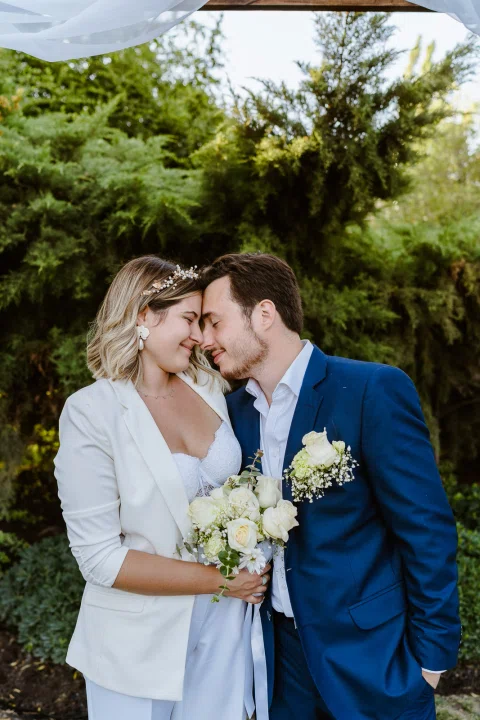 Mujer vestida de blanco y hombre con traje azul juntan sus frentes en un jardín.