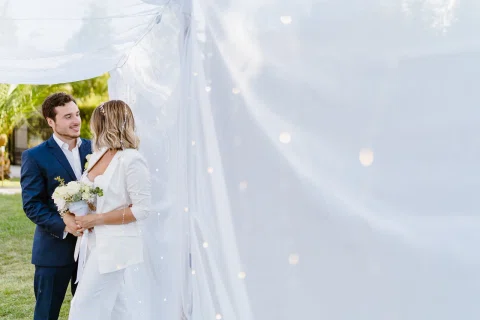 Mujer vestida de blanco con un ramo y hombre con traje azul están juntos entre telas blancas al aire libre.