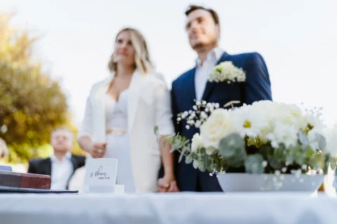 Mujer vestida de blanco y hombre con traje azul están de pie detrás de una mesa con flores en primer plano.