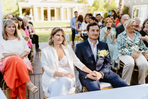 Mujer vestida de blanco y hombre con traje azul están sentados juntos entre otras personas.