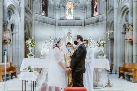 Mujer con vestido blanco y hombre con traje oscuro permanecen tomados de las manos frente a una persona con vestimenta religiosa junto al altar.
