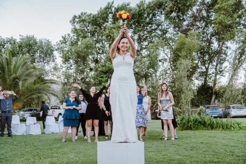 Mujer con vestido blanco levanta un ramo sobre la cabeza frente a un grupo de personas.