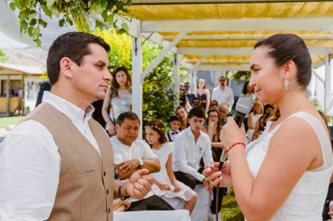 Mujer con vestido blanco sostiene un micrófono frente a un hombre con chaleco beige, con varias personas detrás.