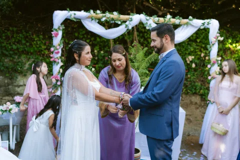 Mujer con vestido morado ata cintas de colores alrededor de las manos unidas de una mujer con vestido blanco y un hombre con traje azul.
