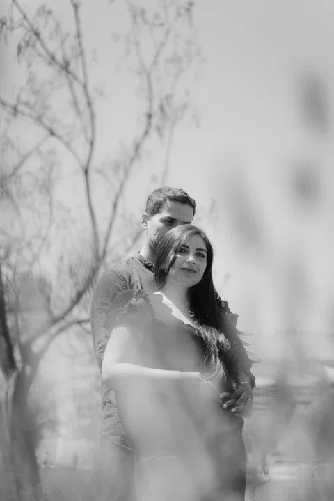 Mujer aparece en primer plano con un hombre detrás, entre ramas desenfocadas, en una fotografía en blanco y negro.