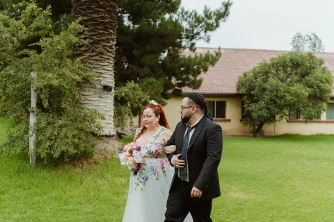 Mujer con ramo y vestido floral camina por el jardín junto a un hombre con traje negro.