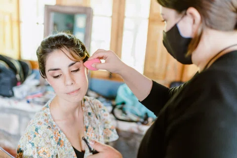Primer plano de una mujer sentada mientras una persona con mascarilla aplica maquillaje en su rostro.