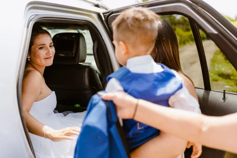 Mujer con vestido blanco está sentada dentro de un automóvil con la puerta abierta, mientras otra persona sostiene a un niño pequeño en el exterior.